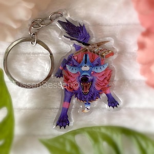 May include: A colorful wolf keychain with a silver keyring. The wolf has a fierce expression with open mouth and sharp teeth. The wolf's fur is a mix of purple, pink, and blue. The keychain is a unique accessory.