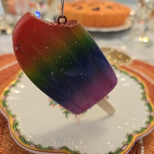 May include: A rainbow-colored popsicle ornament with a wooden stick, hanging from a silver chain. The ornament is resting on a white plate with a floral design and a gold trim. The background includes a pie and other decorative items.