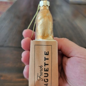 May include: Gold glass bottle ornament with a brown paper label that says "French Baguette".
