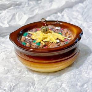 May include: A brown ceramic Christmas ornament shaped like a crock pot filled with a colorful chili-like dish. The dish is topped with a yellow cheese-like substance and red and green sprinkles.