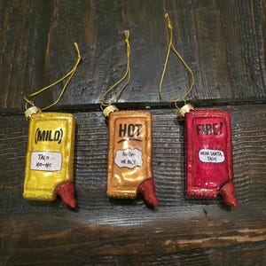 May include: Three glass ornaments shaped like hot sauce bottles. The bottles are decorated with different levels of heat: "(Mild)" with the text "Taco Ho-Ho", "Hot" with the text "Nacho or Nice", and "Fire!" with the text "Dear Santa, Tacos".