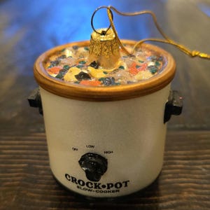 May include: A miniature Crock-Pot slow-cooker Christmas ornament. The white ceramic pot has a brown rim and black handles. The top is filled with colorful, textured food. The text "CROCK-POT SLOW-COOKER" is printed on the front, with settings for "OFF", "LOW", and "HIGH".
