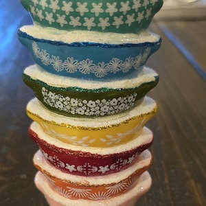 May include: A glass Christmas ornament shaped like a stack of colorful mixing bowls with white floral designs. The bowls are stacked from largest to smallest, with the top bowl being teal, followed by blue, green, yellow, red, orange, and pink.