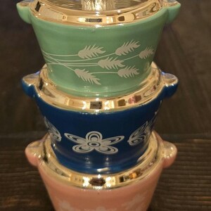 May include: A stack of three glass Christmas ornaments shaped like vintage kitchen mixing bowls. The top bowl is light green with a white wheat design, the middle bowl is blue with a white flower design, and the bottom bowl is pink with a white snowflake design.