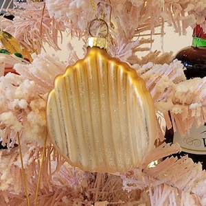 May include: A gold glass ornament shaped like a seashell with a textured surface. The ornament is hanging on a pink artificial Christmas tree.