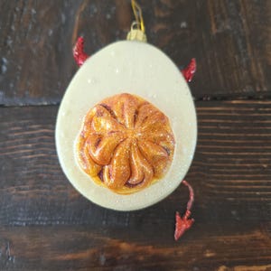 May include: A novelty Christmas ornament shaped like a deviled egg. The egg white is off-white with glitter, and the yolk is a textured orange. Red devil horns and a tail add to the design. A gold-colored loop is at the top for hanging.