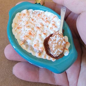 May include: A teal blue ceramic ornament shaped like a casserole dish with a white and brown filling topped with white sprinkles. A small silver spoon is included in the dish.