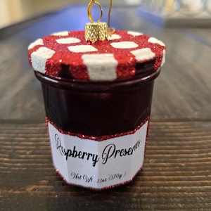 May include: A red and white glass jar ornament with a glittery finish. The jar has a label that reads "Raspberry Preserves Net Wt: 13oz (370g)".