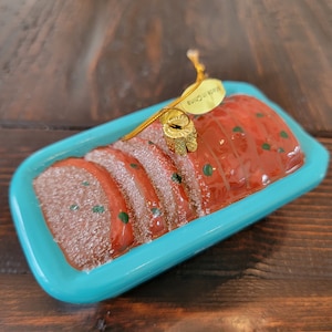 May include: A glass Christmas ornament shaped like a blue casserole dish with a red glazed ham inside. The ornament has a gold string for hanging and a tag that reads "Made in China".