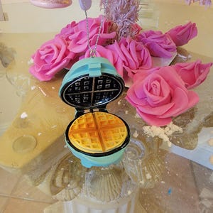 May include: A blue and yellow waffle iron ornament with a waffle design. The ornament is hanging from a silver chain.
