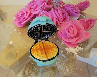 May include: A blue and yellow waffle iron ornament with a waffle design. The ornament is hanging from a silver chain.