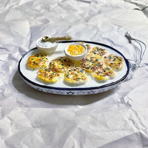May include: A ceramic plate with a white rim and a blue Star of David pattern holds miniature latkes, a small bowl of sour cream, and a bowl of applesauce. A tiny gold spoon rests in the sour cream. The plate is a decorative ornament.