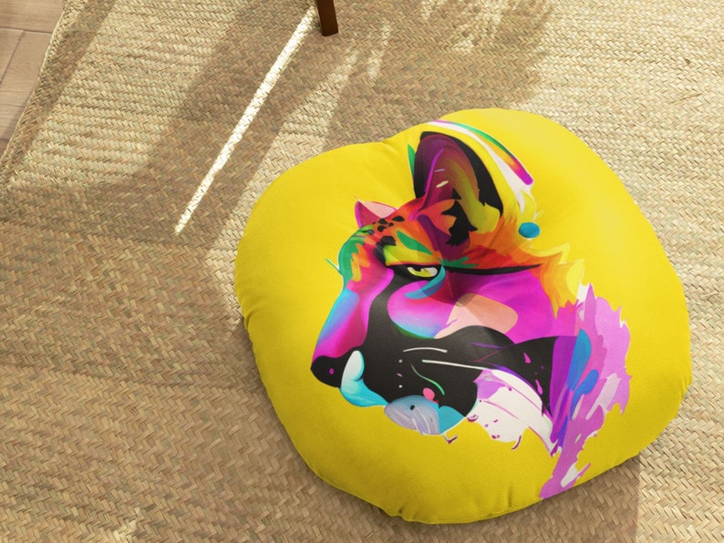 May include: A yellow round floor pillow with a colorful abstract illustration of a cat's face. The cat's fur is pink, orange, blue, and green.