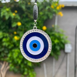 May include: A blue and white evil eye charm with a silver rim and rhinestones. The charm is hanging from a black cord with a black lava stone bead.