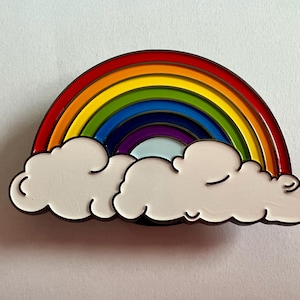 Op de afbeelding: Een kleurrijke emaille pin met een regenboog ontwerp. De regenboog heeft rode, oranje, gele, groene, blauwe, indigo en violette bogen. Twee witte wolken met zwarte contouren bevinden zich onder de regenboog.