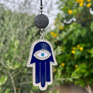 Könnte beinhalten: Ein blauer Hamsa-Hand-Anhänger mit weißem Augendesign und klaren Strass-Akzenten. Der Anhänger hängt an einer schwarzen Kordel mit einer porösen, dunkelgrauen Lavasteinperle. Der Hintergrund ist verschwommenes grünes Laub.