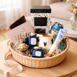 May include: A wicker basket containing gifts such as a candle, blue bottles, a can of soda, a fan, and a card reading "Mom". The basket is on a wooden table, with a ribbon and decorative paper.