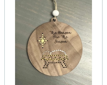 May include: Round wooden Christmas ornament with the text "The Reason For The Season". It features a star and a manger scene. The ornament is light brown and has a string for hanging. The diameter is approximately 8 cm.