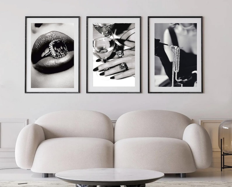 May include: A triptych of framed black and white photographs. One shows a close-up of lips with a ring, another a hand with rings and a glass, and the third a person holding a pearl necklace. The images are displayed above a cream-colored sofa.