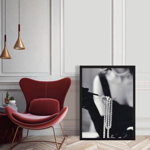 May include: Black and white photograph of a woman wearing a black dress and gloves, holding a strand of pearls. The photo is framed in a black frame.