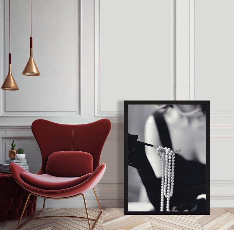 May include: Black and white photograph of a woman wearing a black dress and gloves, holding a strand of pearls. The photo is framed in a black frame.