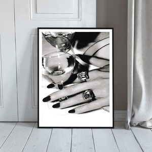 May include: Black and white framed print featuring a close-up of a hand adorned with large, ornate rings, holding a wine glass. The nails are painted black. The image evokes luxury and elegance.