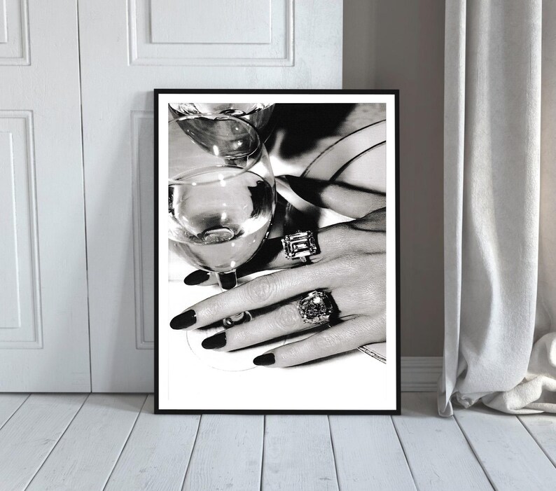 May include: Black and white framed print featuring a close-up of a hand adorned with large, ornate rings, holding a wine glass. The nails are painted black. The image evokes luxury and elegance.