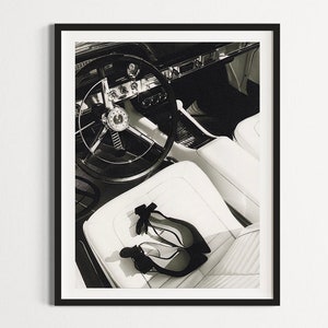 May include: Black and white photo of the interior of a vintage car with a steering wheel and two pairs of black heels on the white leather seat.
