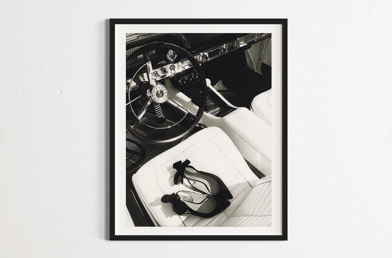 May include: Black and white photo of the interior of a vintage car with a steering wheel and two pairs of black heels on the white leather seat.