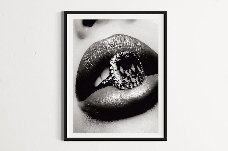 May include: Black and white framed print featuring a close-up of glossy lips with a large diamond ring. The ring has a large, dark-colored center stone surrounded by smaller, clear stones. The image is a high-contrast, artistic photograph.