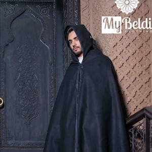 May include: A black hooded cloak with a long, flowing design. The cloak is worn by a person standing in front of a wooden door with intricate carvings.