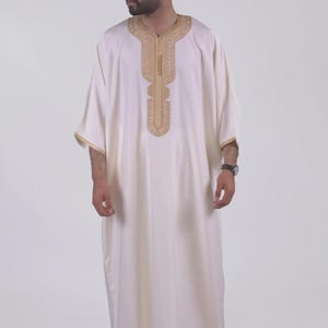 May include: Off-white, long-sleeved garment with gold embroidery around the neckline and down the front. The sleeves have gold trim. The garment is worn with matching off-white shoes. The style is reminiscent of traditional Middle Eastern attire.