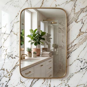 May include: A rectangular mirror with a gold frame reflects a bathroom interior. The vanity is white, and there are plants in terracotta pots. The marble wall and gold fixtures create a luxurious feel. The mirror has rounded corners.