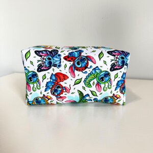 May include: A rectangular zippered pouch with a white background featuring a repeating pattern of blue alien characters dressed as various insects, including butterflies, ladybugs, and caterpillars, interspersed with small green leaves.