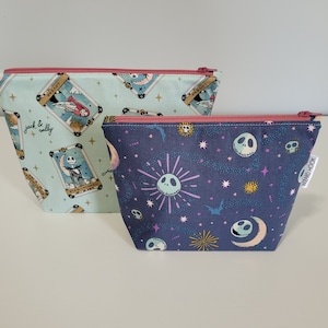 May include: Two zippered pouches with a colorful print. The first pouch is light blue with a white and gold print of a tarot card with Jack and Sally from The Nightmare Before Christmas. The second pouch is dark blue with a white, pink, and yellow print of Jack Skellington's face and stars. The text "WENDYBROOK" is printed on the side of the second pouch.