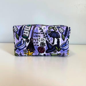 May include: A rectangular zippered pouch featuring a light purple and white gingham pattern with various illustrations including a broom, a witch hat, an umbrella, flowers, and the text "There's a little witch in all of us" and "You were always on my mind."