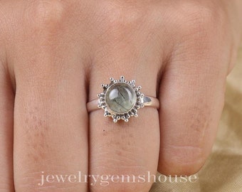 May include: A silver ring featuring a round, translucent green rutilated quartz gemstone with visible dark needle-like inclusions. The stone is set in a sunburst-style bezel with small silver beads radiating outward along the circular edge.