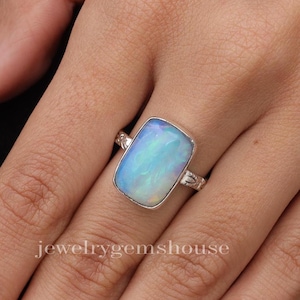 Puede incluir: Un anillo de plata con una piedra de ópalo rectangular. El ópalo es de color azul claro con destellos blancos e iridiscentes.