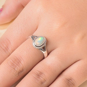 May include: A silver ring featuring an oval opal gemstone. The ring has a delicate design with a split band and a beaded edge around the stone.