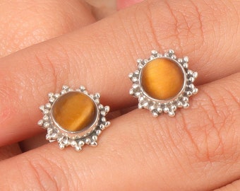 May include: A pair of stud earrings featuring a round, golden-brown tiger's eye stone set in a silver-colored, sunburst-style frame. The earrings are displayed on a light-colored surface.