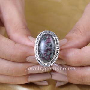 Puede incluir: Un anillo de plata con una piedra ovalada verde y rosa engastada en un engaste de plata detallado. El anillo está en una mano.