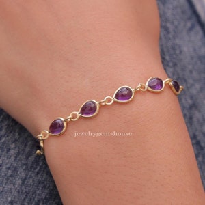 May include: A gold bracelet featuring teardrop-shaped purple gemstones. The gemstones are set in individual bezels and linked together with small gold connectors. The bracelet is shown on a wrist, with the jewelrygems house logo visible.