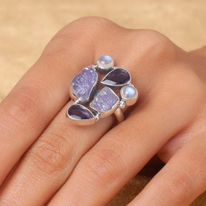 Könnte beinhalten: Ein silberner Ring mit einem Cluster aus violetten und weißen Edelsteinen. Der Ring verfügt über eine Vielzahl von Steinformen und -größen, darunter runde, ovale und unregelmäßige Formen.