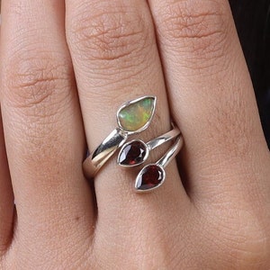 Puede incluir: Un anillo de plata con una ópalo en forma de pera y dos granates en forma de lágrima. El diseño del anillo se envuelve alrededor del dedo, mostrando las piedras preciosas. El ópalo tiene un tono verde pálido, mientras que los granates son de un rojo intenso.