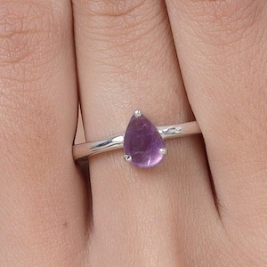May include: A silver ring featuring a pear-shaped, purple amethyst gemstone. The ring's band is thin and simple, with the gemstone held in place by small prongs. The amethyst has a smooth, polished surface.