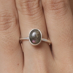 Puede incluir: Un anillo de plata con una piedra ovalada de color gris oscuro. La piedra tiene un sutil brillo iridiscente.
