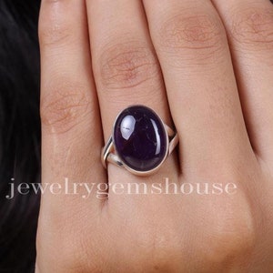 May include: A silver ring with a large, oval amethyst gemstone. The dark purple stone is set in a simple band. The ring is displayed on a finger. The text "jewelrygemshouse" is visible.