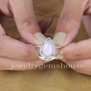 May include: A silver ring featuring a teardrop-shaped pink gemstone set in a beaded bezel. The ring is worn on a finger.