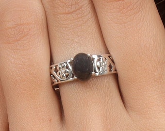 Könnte beinhalten: Ein silberner Ring mit einem ovalen, dunkelgrauen Stein. Das Band hat ein filigranes, offenes Design mit floralen und geometrischen Mustern. Der Ring wird am Finger getragen.