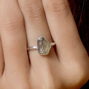 Puede incluir: Un anillo de plata con una piedra preciosa verde pálido de forma irregular. El anillo tiene una banda fina y pulida, y la piedra preciosa está engastada en un estilo simple y minimalista. El anillo se muestra en un dedo.